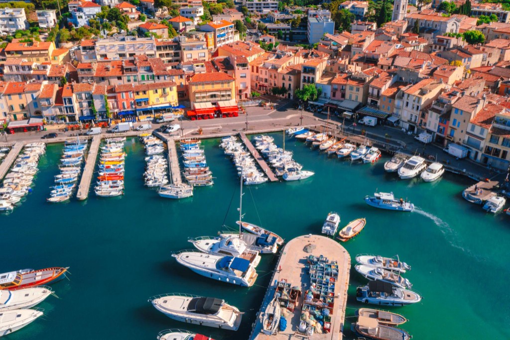 Aerial of a Mediterranean harbor: boats packed in a marina, a tight quay, and a continuous row of shops and houses